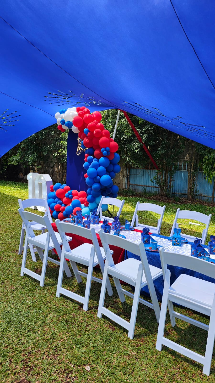 A blue and red birthday table with white chairs and a matching balloon display under a canopy