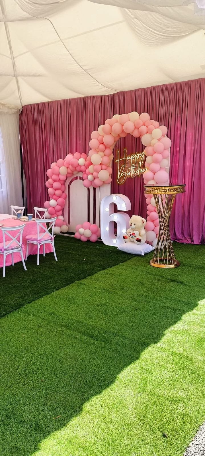 A pink sixth birthday setup with balloon arches, an illuminated number and a dressed children's table