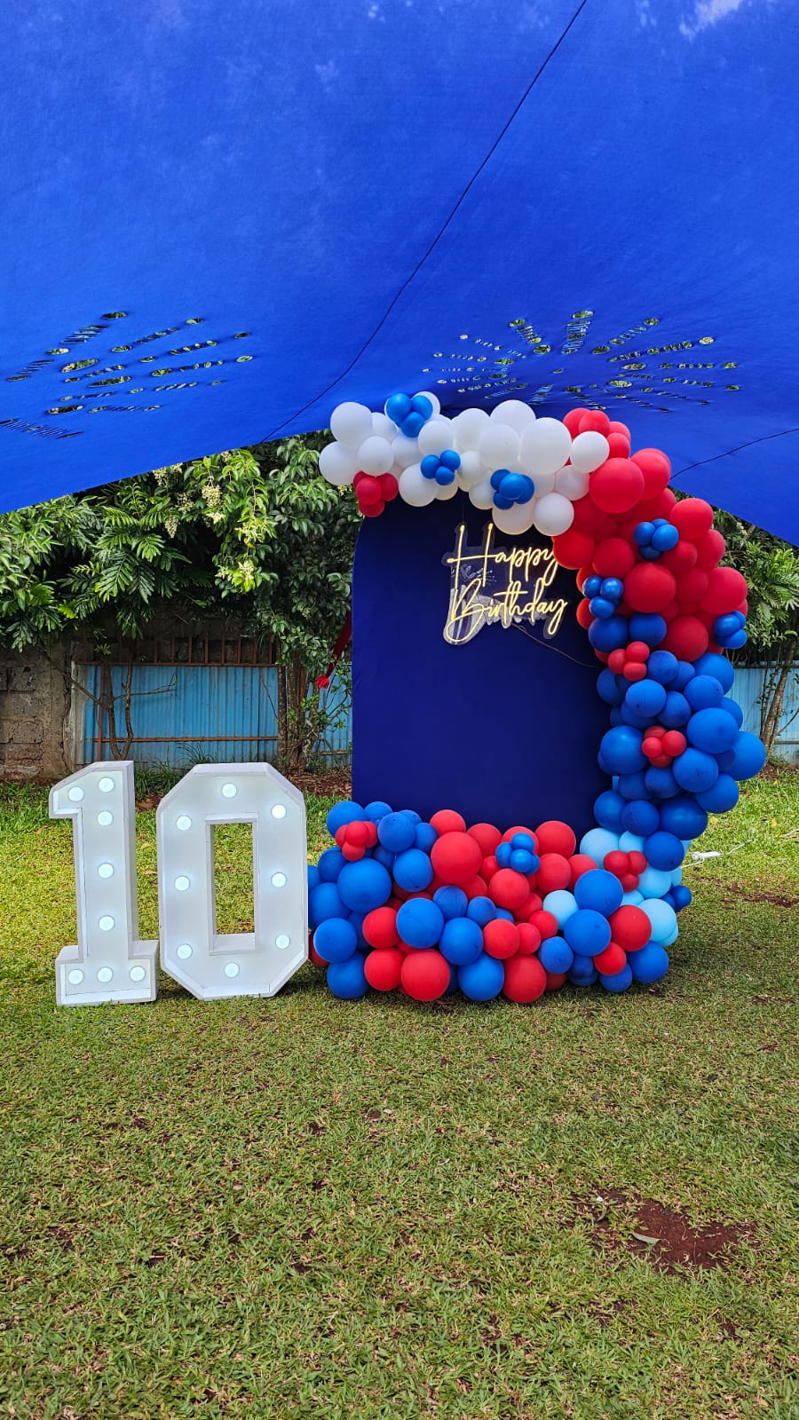 A blue, red and white balloon birthday backdrop with an illuminated number display on the lawn