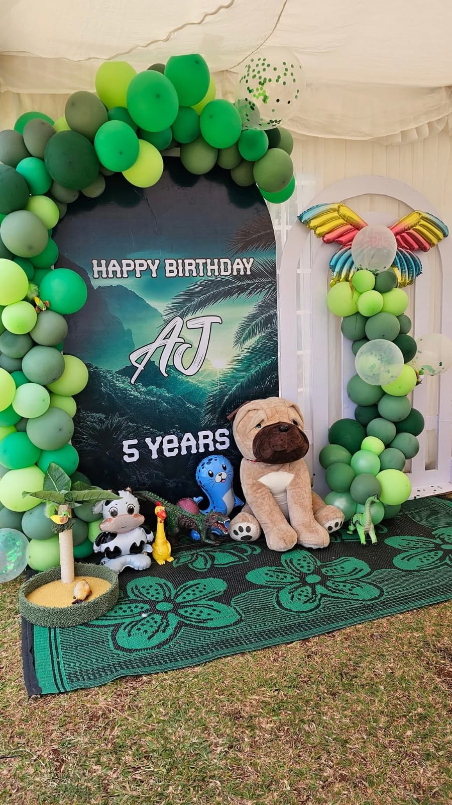 A green animal-themed fifth birthday backdrop with balloons and plush animal decorations